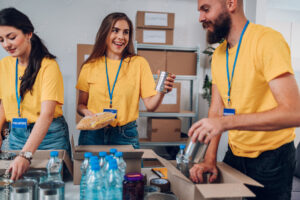 volunteers in yellow t-shirts wearing blue badges on blue lanyards packing food boxes, a woman is smiling a a man holding a bag of noodles and a can of food, the man is putting cans into a cardboard box