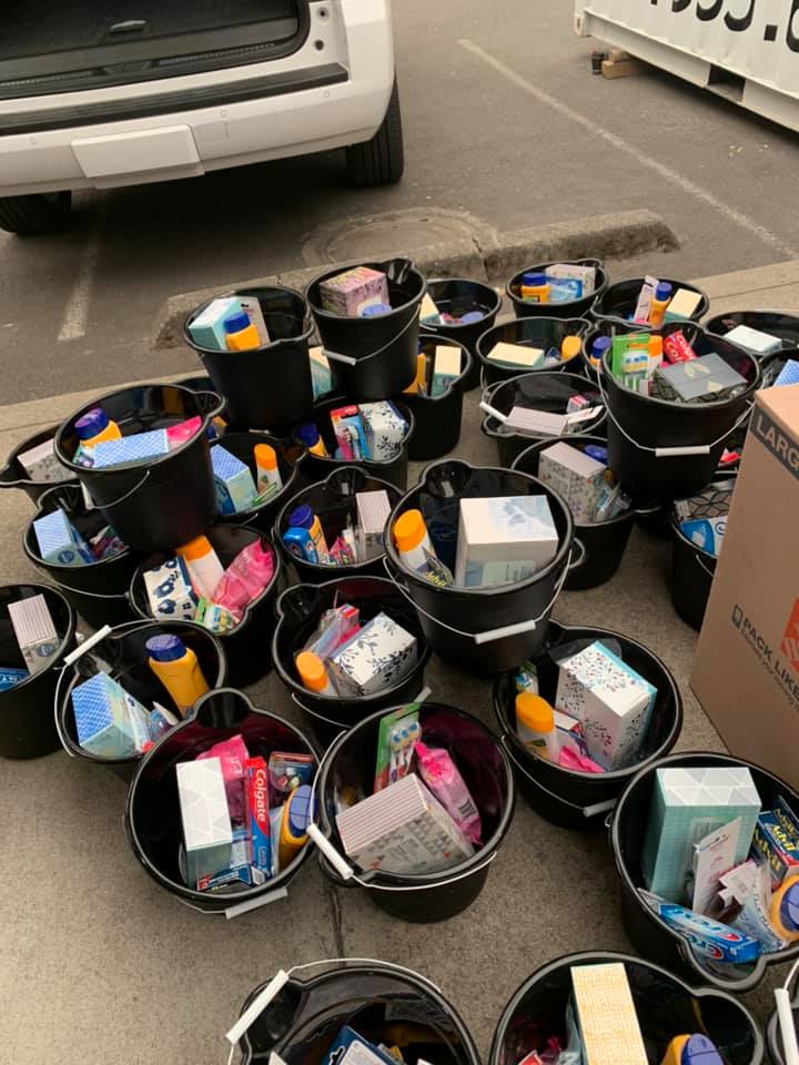 Lots of black buckets on the ground filled with toiletries items including toothpaste, kleenex, q-tips, toothbrushes, and more.