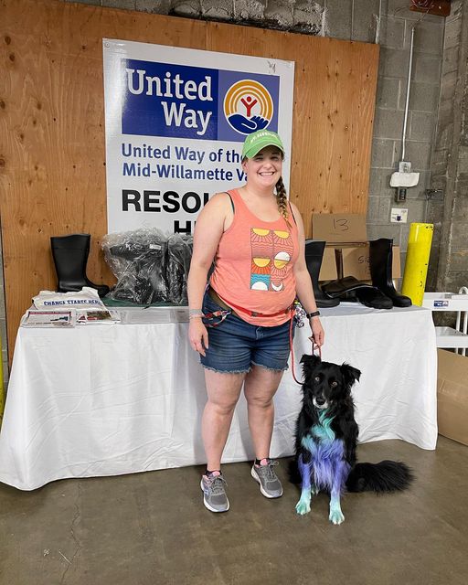 A girl wearing a salmon colored tank top, shorts, and green hat stands with her dog on a leash in front of the United Way of the Mid-Willamette Valley Good 360 boot table
