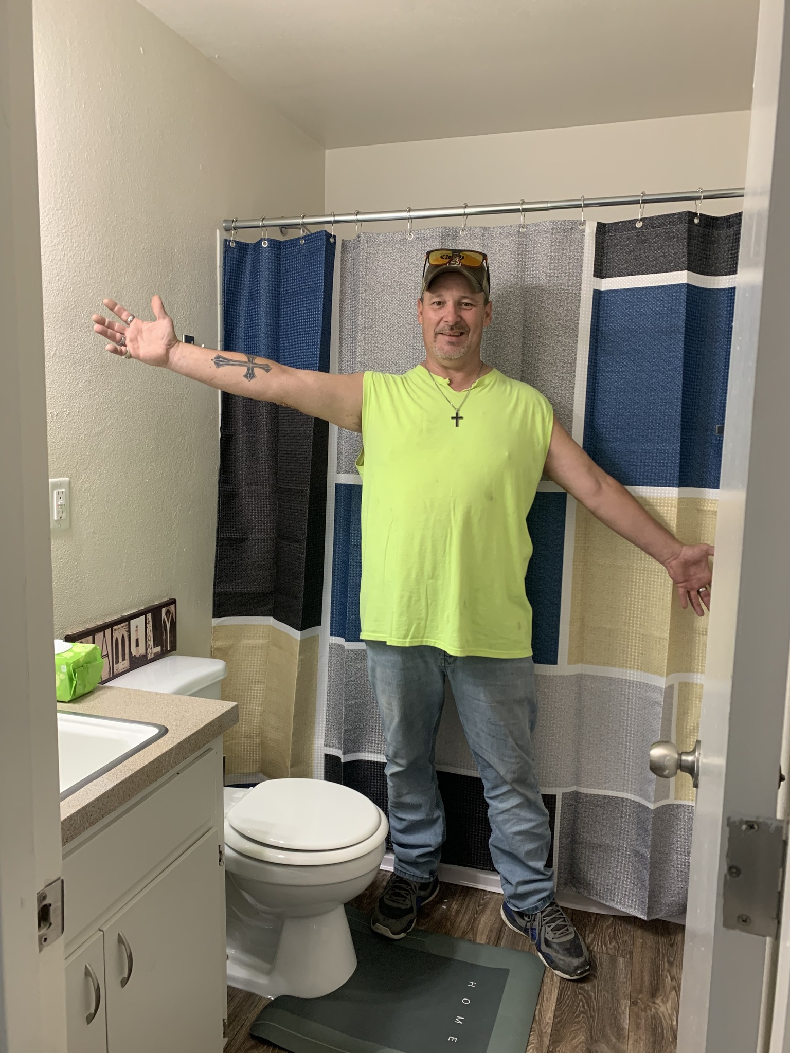 A man standing in a bathroom with his arms outstretched, wearing a yellow shirt.
