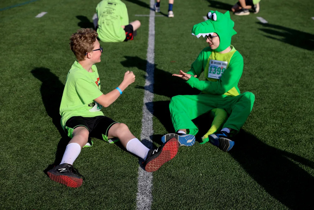 one boy wearing a green shirt and a kid in an alligator costume playing rock, paper, scissors sitting on a field