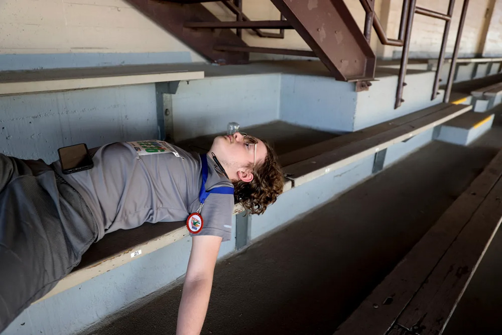 a teenage boy laying on a bleacher resting wearing a gray tshirt and phone on his stomach