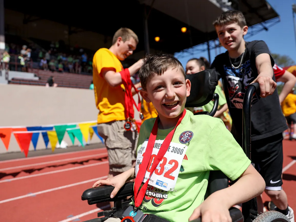 a boy in a wheelchair wearing a green t-shirt smiling with his medal being pushed by a boy wearing all black guy in yellow handing out medals