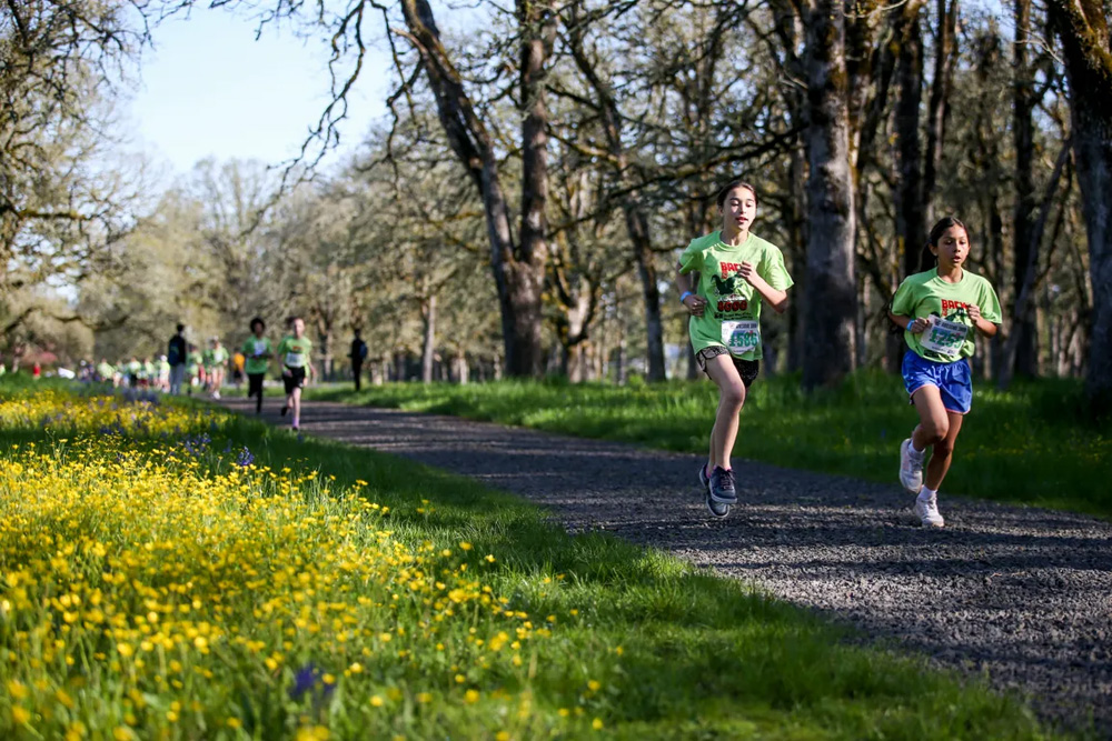 two girls in green t-shirts running on a gravel path outside with green grass. yellow flowers, and trees